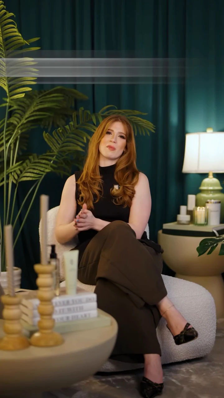 A red-haired presenter seated in a cream chair against green curtains